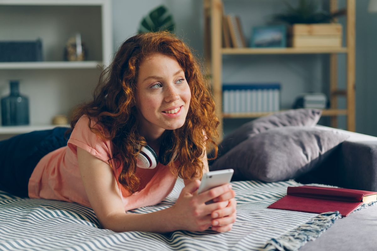 Redheaded woman smiling while using a smartphone, lying on bed at cozy home.